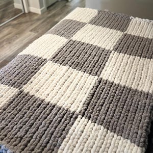 May include: A chunky knit blanket in a checkerboard pattern. The blanket features alternating squares of gray and off-white. The thick yarn creates a textured, cozy appearance, perfect for adding warmth and style to any space.
