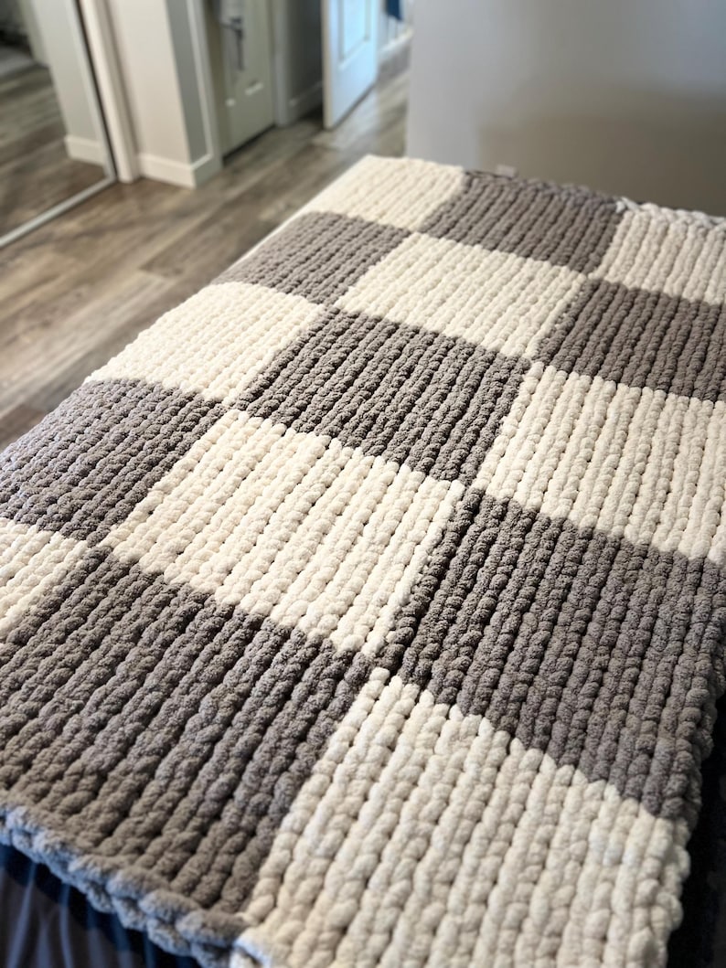 May include: A chunky knit blanket in a checkerboard pattern. The blanket features alternating squares of gray and off-white. The thick yarn creates a textured, cozy appearance, perfect for adding warmth and style to any space.