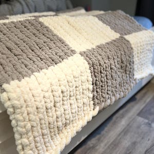 May include: A chunky knit blanket in a checkerboard pattern. The blanket features alternating squares of light gray and cream-colored yarn. The thick yarn creates a textured, cozy appearance, perfect for home decor.