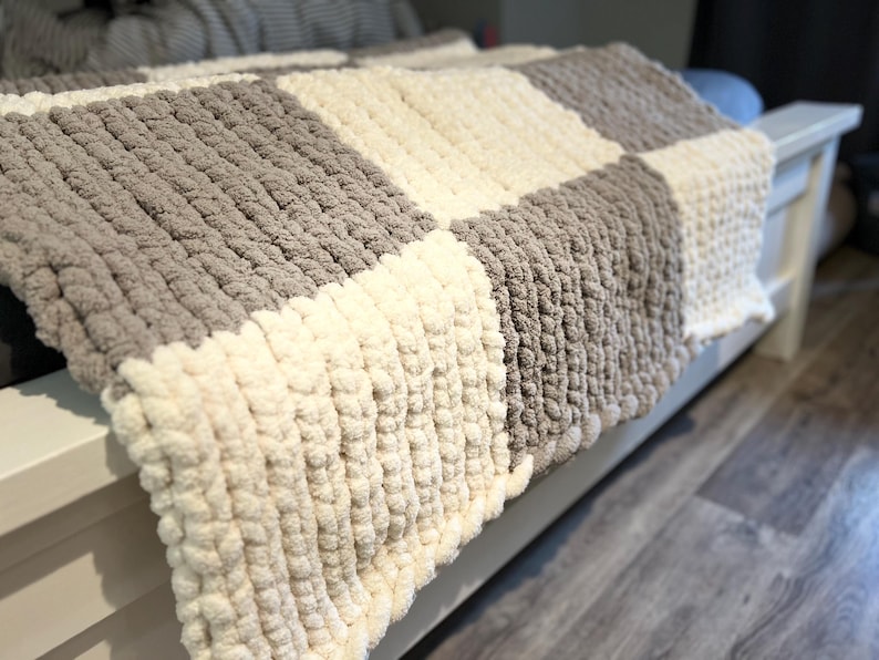 May include: A chunky knit blanket in a checkerboard pattern. The blanket features alternating squares of light gray and cream-colored yarn. The thick yarn creates a textured, cozy appearance, perfect for home decor.