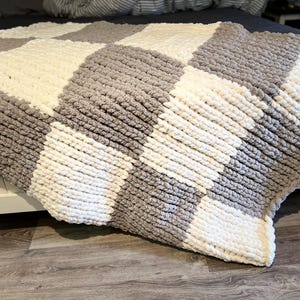 May include: A chunky knit blanket in a checkerboard pattern of gray and off-white squares. The blanket is draped over a bed, showcasing its soft texture and cozy design. This handmade blanket is perfect for adding warmth and style to any bedroom.