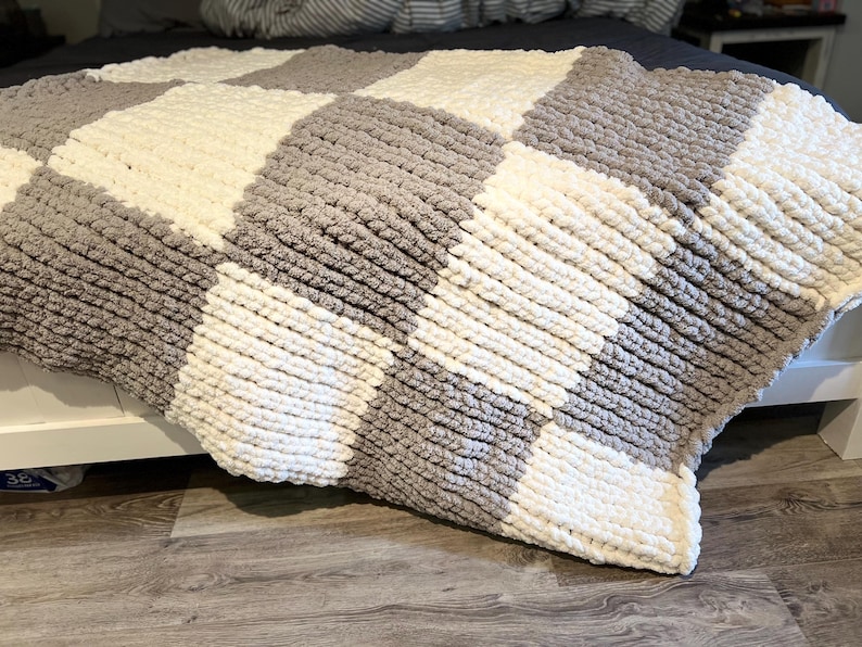 May include: A chunky knit blanket in a checkerboard pattern of gray and off-white squares. The blanket is draped over a bed, showcasing its soft texture and cozy design. This handmade blanket is perfect for adding warmth and style to any bedroom.