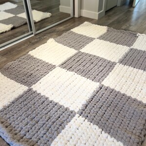 May include: A handmade, checkerboard-patterned rug in shades of gray and white. The rug is made of a thick, plush material, with a soft, textured appearance. The design features alternating squares of gray and white, creating a classic, modern look.
