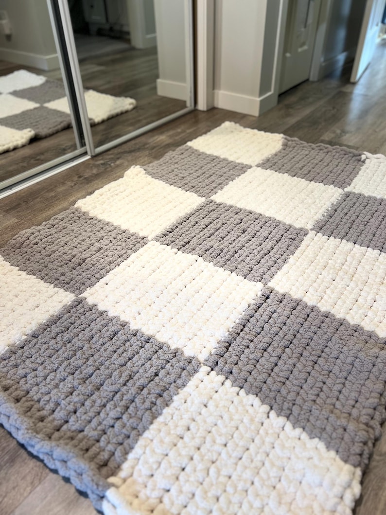 May include: A handmade, checkerboard-patterned rug in shades of gray and white. The rug is made of a thick, plush material, with a soft, textured appearance. The design features alternating squares of gray and white, creating a classic, modern look.