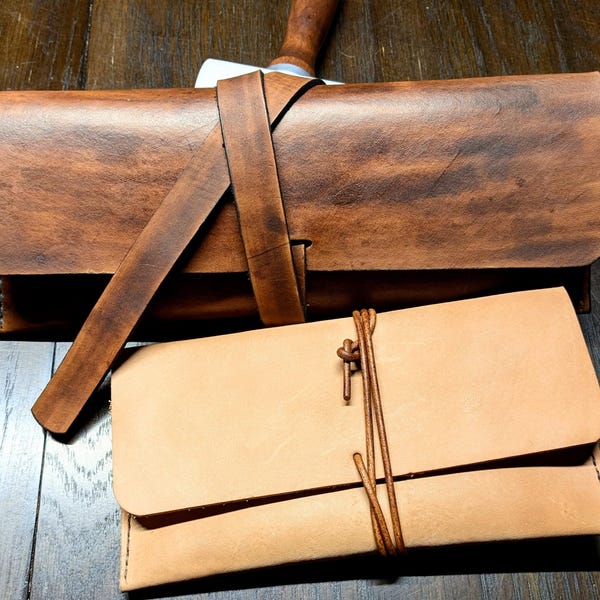May include: Two rectangular leather cases. The top case is dark brown with a wide leather strap closure. The bottom case is light tan with a thin leather cord wrap closure. Both cases feature a flap design and visible stitching along the edges.