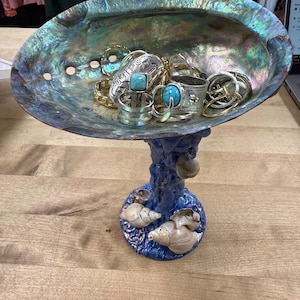 May include: An iridescent abalone shell bowl mounted on a blue textured pedestal base decorated with small seashells. The bowl contains a collection of various metal rings, including two silver-toned rings featuring inlaid turquoise-colored stones.