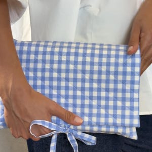 Könnte beinhalten: Eine rechteckige, gesteppte Clutch mit einem hellblau-weißen Vichy-Karo-Muster. Das Stoffaccessoire verfügt über ein passendes, am vorderen Umschlag befestigtes Vichy-Band zum Binden, das als Verschluss für die gefaltete Tasche dient.