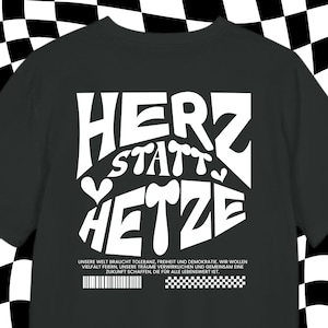 Może przedstawiać: Czarny T-shirt z białym tekstem, który brzmi "HERZ STATT HETZE". Pod tekstem znajduje się mniejszy biały tekst, który brzmi "UNSERE WELT BRAUCHT TOLERANZ, FREIHEIT UND DEMOKRATIE. WIR WOLLEN VIELFALT FEIERN, UNSERE TRAUME VERWIRKLICHEN UND GEMEINSAM EINE ZUKUNFT SCHAFFEN, DIE FÜR ALLE LEBENSWERT IST."