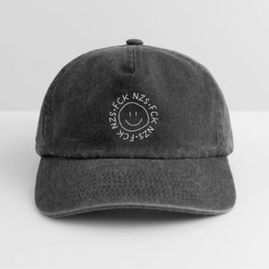 Könnte beinhalten: Eine anthrazitfarbene Baseballkappe mit fünf Paneelen und gebogenem Schirm. Mittig auf der Vorderseite befindet sich eine weiße Stickerei mit einem handgezeichneten Smiley in einem Kreis, der den Text FCK NZS umschließt.