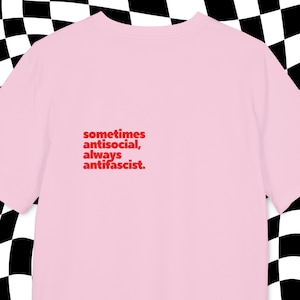 May include: A pale pink t-shirt featuring the red text: "sometimes antisocial, always antifascist". The shirt is made of a soft fabric, suitable for casual wear. The background has a black and white checkered pattern.