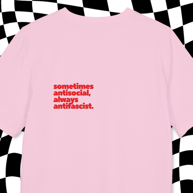 May include: A pale pink t-shirt featuring the red text: "sometimes antisocial, always antifascist". The shirt is made of a soft fabric, suitable for casual wear. The background has a black and white checkered pattern.