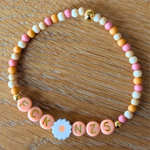 Könnte beinhalten: Ein farbenfrohes Perlenarmband mit pfirsichfarbenen, weißen, rosa und orangefarbenen Perlen. Das Armband hat pfirsichfarbene Buchstabenperlen, die ein Wort bilden, und eine weiße blütenförmige Perle. Goldfarbene Perlen sind eingestreut.