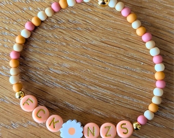 May include: A beaded bracelet with a mix of peach, white, pink, and orange beads. The bracelet features peach-colored letter beads spelling out a word, and a white daisy-shaped bead. Gold-colored beads are interspersed throughout.