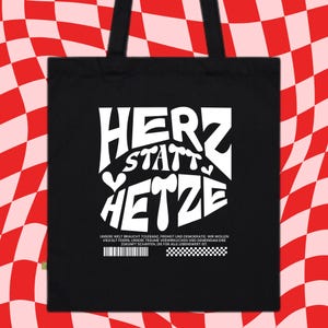 May include: Black tote bag with white text that reads "Herz statt Hetze" which translates to "Heart instead of Hate". The text is in a bold, stylized font. Below the text is a smaller line of text that reads "Unsere Welt braucht Toleranz, Freiheit und Demokratie. Wir wollen Vielfalt feiern, unsere Träume verwirklichen und gemeinsam eine Zukunft schaffen, die für alle lebenswert ist." which translates to "Our world needs tolerance, freedom and democracy. We want to celebrate diversity, realize our dreams and together create a future that is worth living for all."
