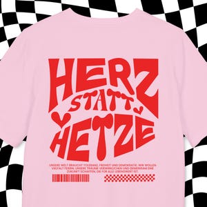 Op de afbeelding: Een lichtroze t-shirt met de Duitse woorden "HERZ statt HETZE" in het rood, wat "Hart in plaats van Haat" betekent. Het shirt heeft ook een verklaring in het Duits onderaan. De achtergrond heeft een zwart-wit geblokt patroon.