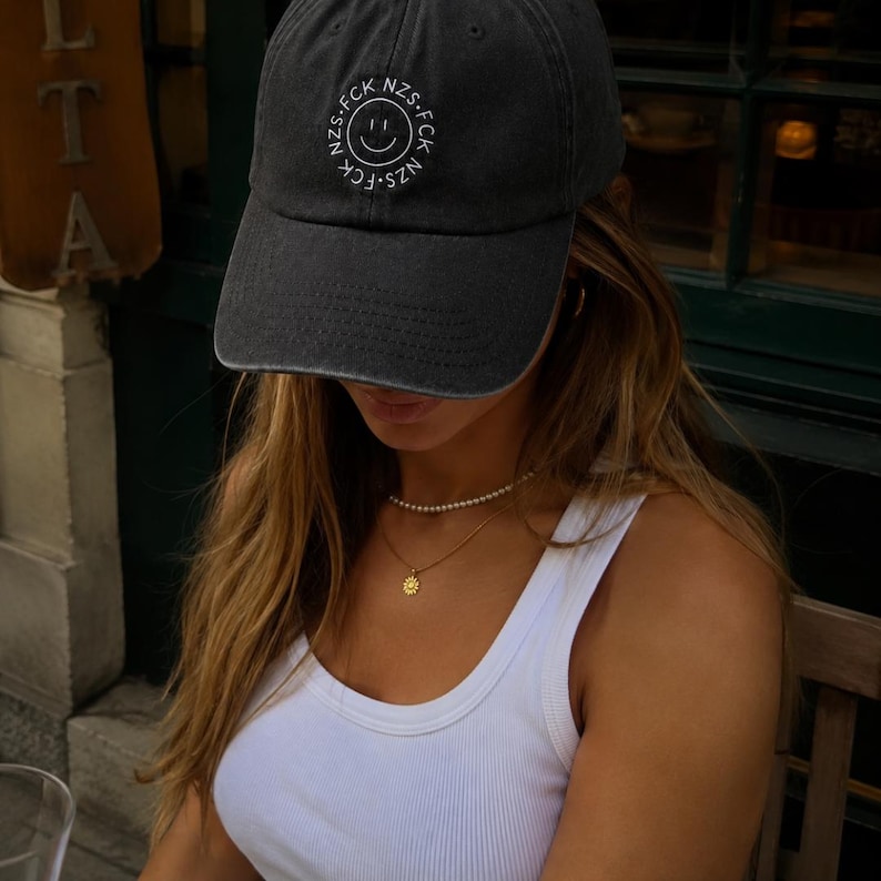 May include: A charcoal gray baseball cap featuring a white embroidered circular design on the front. The design consists of a central smiley face surrounded by the repeating text FCK NZS in a circular pattern. The cap has a curved brim.