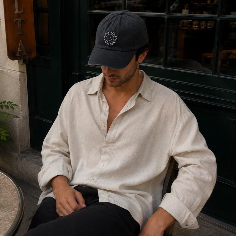 May include: A charcoal gray baseball cap featuring a circular white embroidered logo with a smiley face and the text NZS-FCK NZS-FCK, paired with a long-sleeved, button-down shirt in a light beige, textured fabric and solid black trousers.
