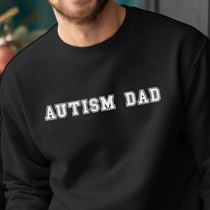 Peut inclure: Sweat-shirt noir à col rond avec l'inscription "AUTISM DAD" en lettres blanches. Le sweat-shirt est de couleur unie et semble être fait d'une matière douce. Le texte est centré sur la poitrine.