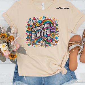 May include: A soft cream colored t-shirt with a colorful graphic design that says "Neurodiversity is Beautiful". The graphic features a rainbow, flowers, and puzzle pieces.