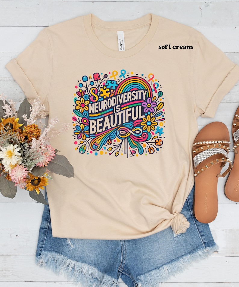 May include: A soft cream colored t-shirt with a colorful graphic design that says "Neurodiversity is Beautiful". The graphic features a rainbow, flowers, and puzzle pieces.