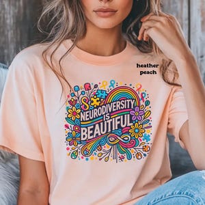 Neurodiversity is beautiful is shown through the print and is the colour of the shirt. It is outlined in black. On this heather peach shirt, vibrant flowers, autism symbols and puzzle pieces surround the words.