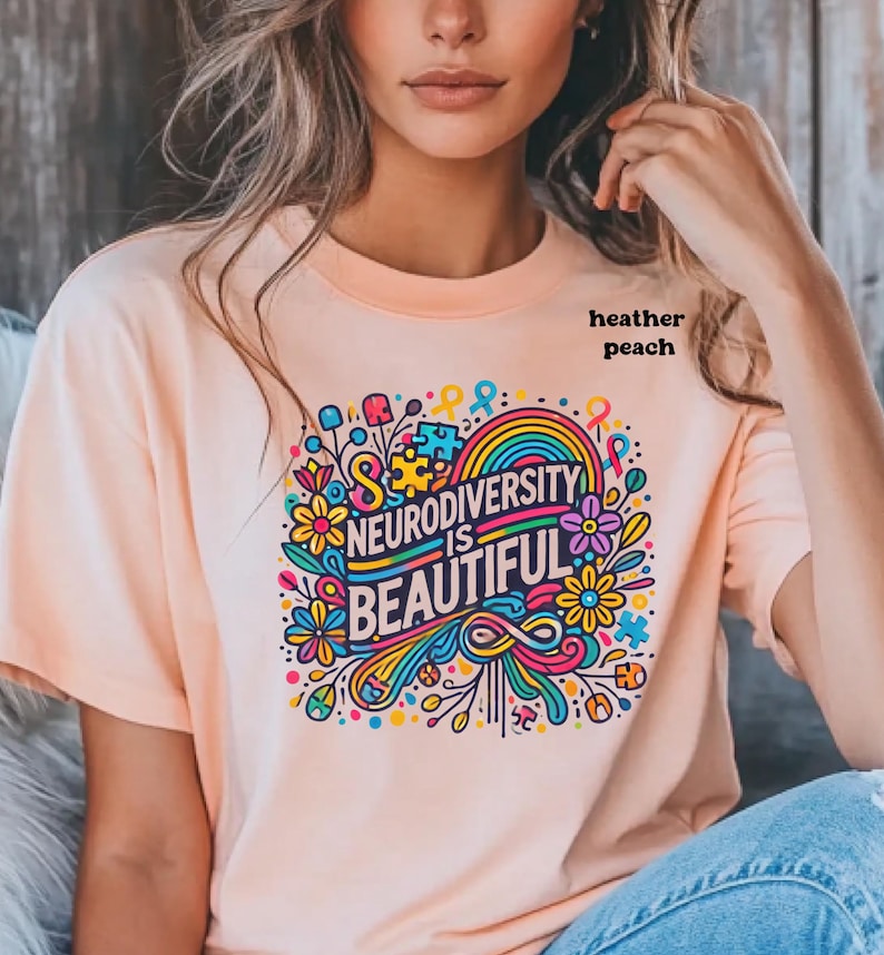 Neurodiversity is beautiful is shown through the print and is the colour of the shirt. It is outlined in black. On this heather peach shirt, vibrant flowers, autism symbols and puzzle pieces surround the words.