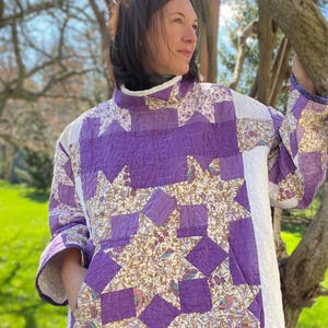 May include: A quilted jacket featuring a star pattern in shades of purple, white, and floral print fabric. The jacket has a high collar and long sleeves. The design incorporates a mix of textures and patterns, creating a unique and artistic look.