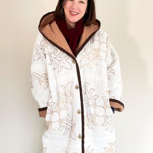 May include: White quilted coat with a floral design in gold stitching. The coat has a brown trim, a hood, and is buttoned up. The lining is a light brown color. The person is wearing a burgundy scarf and blue jeans.