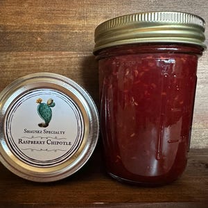 Può includere: Un barattolo di vetro pieno di marmellata di lamponi chipotle rossa. Il barattolo ha un coperchio dorato e un'etichetta bianca con un'illustrazione di cactus verde e il testo "Shausez Specialty Raspberry Chipotle".