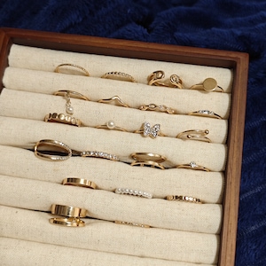 May include: A wooden jewellery box displaying an assortment of gold-coloured rings. The rings showcase diverse designs, including bands, delicate chains, and rings embellished with pearls and crystals. The box is lined with cream-coloured fabric.