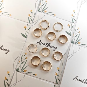 May include: A set of nine gold-toned rings, some with a twisted design, displayed on a white card with floral accents and the text "Avathing".
