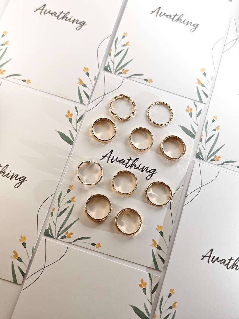 May include: A set of nine gold-toned rings, some with a twisted design, displayed on a white card with floral accents and the text "Avathing".
