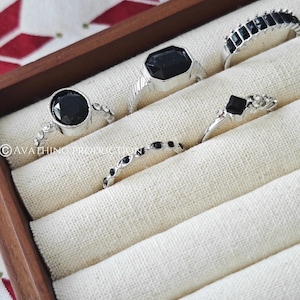 May include: A collection of silver rings with black gemstones displayed in a wooden jewelry box. The rings feature various designs, including round, square, and rectangular stones. The box is lined with a cream-colored fabric.