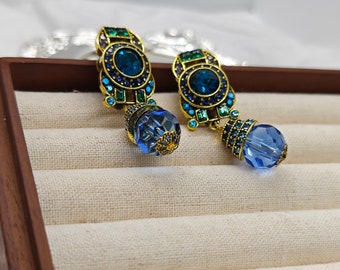 May include: A pair of gold tone dangle earrings with blue and green crystal accents. The earrings feature a round, faceted blue crystal drop.