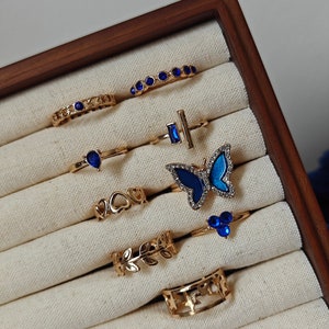 Pode incluir: Uma coleção de anéis dourados expostos numa caixa de joias de madeira. Os anéis apresentam vários designs, incluindo pedras preciosas azuis, uma borboleta, corações, folhas e elos de corrente. Os anéis são colocados sobre um tecido creme.