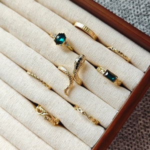 May include: A set of seven gold rings in a wooden box. The rings feature different designs, including a snake, a geometric pattern, and a ring with a green gemstone.