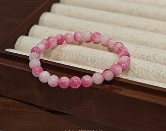 Puede incluir: Un brazalete de cuentas rosa y blanco con un sutil efecto degradado. El brazalete se muestra sobre una superficie de madera con un forro de tela blanco.