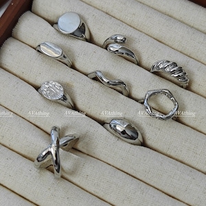 May include: A set of nine silver rings displayed in a wooden box with white fabric lining. The rings are of various shapes and sizes, including a circle, a wave, a rectangle, and a cross.