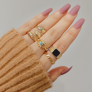 May include: A hand adorned with multiple gold rings. The rings include a twisted band, an eye design with a turquoise stone, a black rectangular signet ring, and a ring with the letter "S". The hand is wearing a brown sweater and has long, matte pink nails.