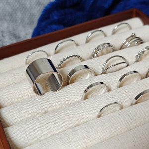 May include: A wooden ring box with a white fabric interior. The box is filled with silver rings of various styles and sizes.