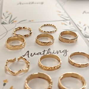 May include: A set of nine gold-toned rings with different designs, including twisted, textured, and open-work styles. The rings are displayed on a white background with the word "Awathing" printed in black.