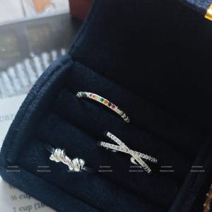 Puede incluir: Una colección de anillos de plata exhibidos en una caja de joyería negra abierta. Los anillos incluyen una banda curva con gemas coloridas, un diseño cruzado con piedras claras y un estilo anudado. El interior oscuro resalta las joyas.