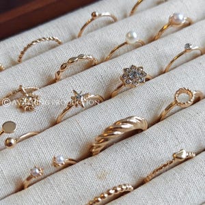 May include: A selection of gold rings in a jewellery box. The rings have diverse designs, including pearls, gemstones, and textured bands. They are arranged on a light fabric, highlighting their intricate details. The rings are in a wooden box.