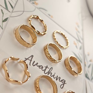 May include: Set of seven gold-toned rings with different designs, including a wavy ring, a ring with a textured surface, and a ring with a braided design. The rings are displayed on a white background with the word "Ava thing" printed on the card.