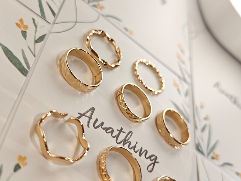 May include: Set of seven gold-toned rings with different designs, including a wavy ring, a ring with a textured surface, and a ring with a braided design. The rings are displayed on a white background with the word "Ava thing" printed on the card.