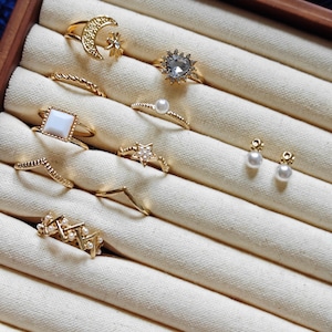 Può includere: Una collezione di anelli e orecchini dorati esposti in un portagioie di legno. Gli anelli presentano vari design, tra cui una luna crescente con una stella, una pietra a forma di cuore e delicate fasce con perle e forme geometriche. Gli orecchini sono piccoli, con borchie di perle.