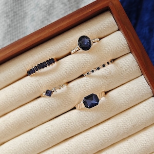 May include: Five gold rings with dark blue gemstones in a wooden ring box. The rings are arranged in a row, with the largest ring at the top and the smallest ring at the bottom.
