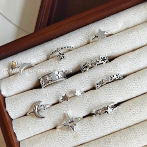 Puede incluir: Una colección de anillos de plata con diseños celestiales, incluyendo estrellas y lunas crecientes. Los anillos se exhiben en una caja de joyería de madera con un forro de tela color crema. Varios anillos en forma de estrella y un anillo con una estrella colgante son visibles.