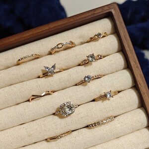 May include: A set of ten gold rings with clear rhinestones, displayed in a wooden ring box with a beige fabric lining.