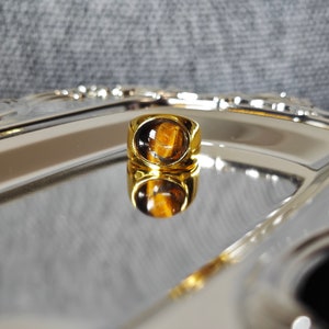 May include: A gold-colored ring with a large, oval tiger's eye stone. The ring is set on a reflective silver tray, creating a mirrored effect. The stone displays bands of brown and gold.